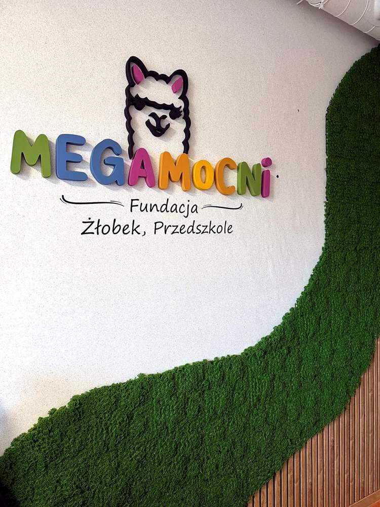 Kolorowy napis MegaMocni z alpaką – Fundacja, Żłobek, Przedszkole – na ścianie z falistą ścianą z zielonego mchu